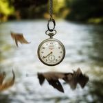 Antique pocket watch suspended in air over a softly blurred river and falling leaves.